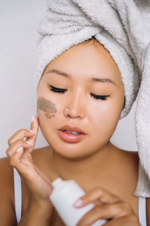 A woman applying facial cream with a white towel wrapped around her hair